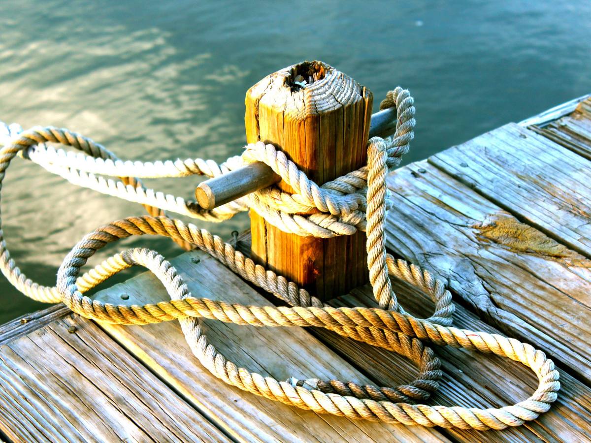 Thick rope looped around a wooden dock post with a metal cleat pin above green water
