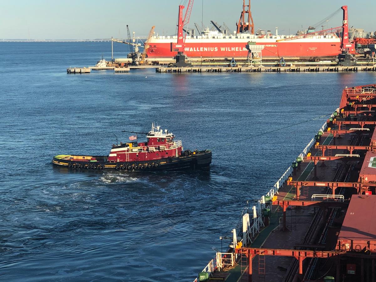 Tugboat working in the Newport News harbor on the James River, with cargo ships and commercial piers behind