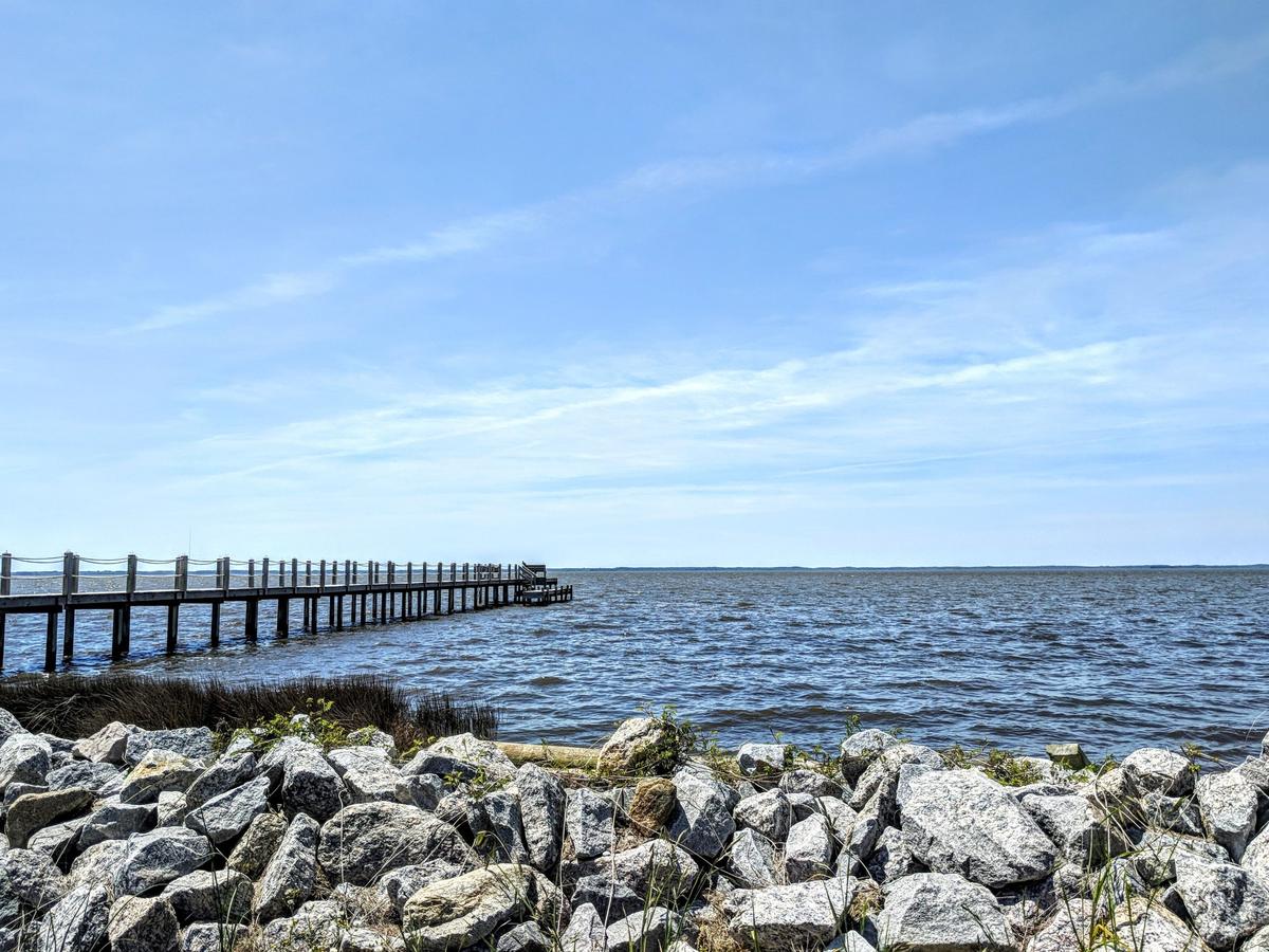 Long wooden pier reaching into open water beside a rip rap shoreline and marsh grass